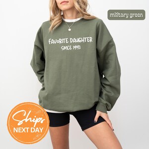 May include: Military green sweatshirt with the text "Favorite Daughter Since 1993" in white. The sweatshirt is a crew neck style and is being worn by a person. The background is plain white.
