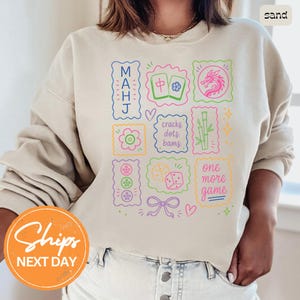 May include: A sand-colored crewneck sweatshirt featuring a colorful graphic print of Mahjong-themed icons, including tiles, a dragon, bamboo, dice, and text that reads MAHJ, cracks dots bams, and one more game, arranged in a grid-like pattern.