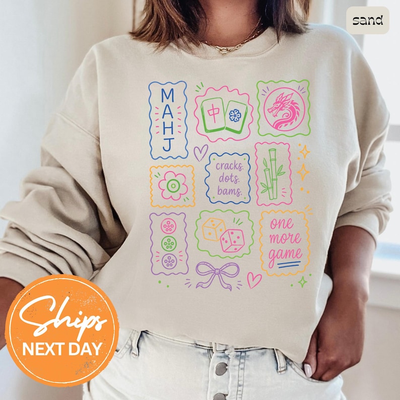 May include: A sand-colored crewneck sweatshirt featuring a colorful graphic print of Mahjong-themed icons, including tiles, a dragon, bamboo, dice, and text that reads MAHJ, cracks dots bams, and one more game, arranged in a grid-like pattern.