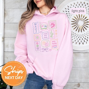 May include: A light pink hooded sweatshirt featuring a graphic print of Mahjong tiles and related symbols. The design includes the text MAHJ, cracks dots bams, and one more game, surrounded by colorful illustrations of tiles, dice, and bamboo.