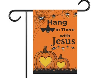 Puede incluir: Una bandera de jardín de Halloween con fondo naranja, con calabazas con recortes de corazón, arañas, un murciélago y el texto "Hang in There with Jesus". La bandera está montada en un soporte de metal negro.