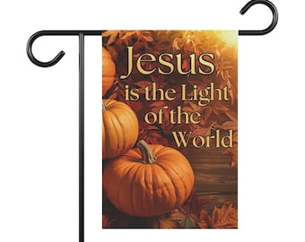 Puede incluir: Una bandera de jardín con el texto dorado "Jesús es la Luz del Mundo". El diseño incluye dos calabazas naranjas y hojas de otoño. La bandera está montada en un soporte de metal negro.