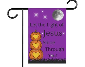 Puede incluir: Una bandera de jardín morada con el texto "Let the Light of Jesus Shine Through You!" presenta tres calabazas naranjas apiladas con recortes de corazón, una luna llena y estrellas. La bandera está montada en un soporte de metal negro.