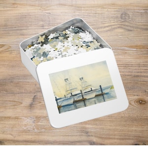 Può includere: Una scatola di puzzle in metallo bianco con un dipinto ad acquerello di barche attraccate a un molo. La scatola è parzialmente aperta, rivelando pezzi di puzzle nei toni del grigio, giallo e bianco. L'immagine è su uno sfondo di legno.