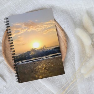 May include: Spiral-bound notebook featuring a vibrant sunset over the ocean. The cover displays a photograph of the sun setting over crashing waves on a sandy beach. The notebook is resting on a wooden tray.