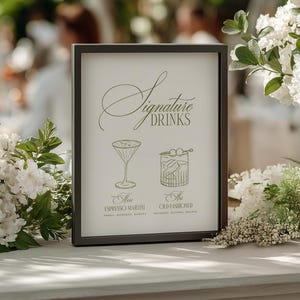 Puede incluir: Un letrero con marco negro con las palabras "Signature Drinks" e ilustraciones de c&oacute;cteles. El letrero est&aacute; sobre una superficie blanca, rodeado de flores blancas y un jarr&oacute;n de rosas blancas. El fondo es borroso con vegetaci&oacute;n.