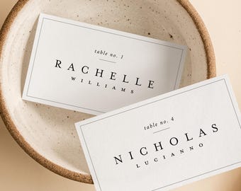 May include: Two white place cards with black text, one reading "RACHELLE WILLIAMS" and the other "NICHOLAS LUCIANNO", are in a speckled ceramic bowl. Dried pampas grass is to the right, all on a beige background.