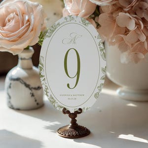Op de afbeelding: Een ovale tafelnummer met het groene nummer 9, geplaatst op een bronzen standaard. De kaart is versierd met een bloemenrand en de namen "Hannah & Matthew" en de datum "06.25.28". Het tafelnummer is omgeven door een bloemstuk.
