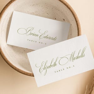 May include: Two white place cards with elegant green calligraphy, one reading "Brian Edwards" and the other "Elizabeth Marshells." They rest in a speckled ceramic bowl, with dried pampas grass on a beige background. Table numbers are also included.