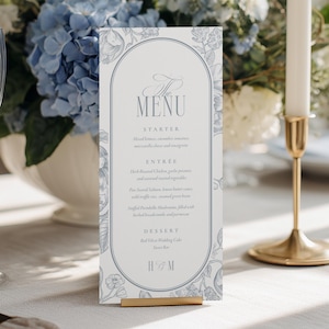 May include: A table setting with a floral centerpiece of blue hydrangeas and white flowers. A menu card with the word "MENU" is displayed. Two wine glasses and two gold candle holders with white candles are also visible.