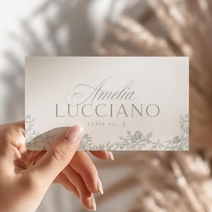 Puede incluir: Una tarjeta rectangular con el nombre "Amelia Lucciano" en escritura elegante y "table no. 2" en letra más pequeña. La tarjeta tiene detalles florales y una paleta de colores suave y neutra, sostenida por una mano.