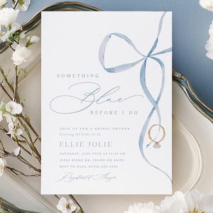 Op de afbeelding: Een uitnodiging voor een bruidsdouche met de tekst "Something Blue Before I Do" en een aquarel blauw lint ontwerp. De uitnodiging staat op een witte kaart, met een illustratie van een gouden ring. Witte bloemen en een decoratieve schaal omringen de kaart.