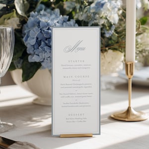 May include: A wedding reception table setting with a menu card, wine glasses, and floral arrangement. The menu card has the word "Menu" at the top. The floral arrangement features blue hydrangeas. Two white candles are in gold candle holders.