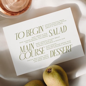 May include: A menu card with a list of courses, a glass of rose wine, and two pears on a white cloth. The menu includes salad, main course, and dessert options. The menu is printed in a serif font with green text.