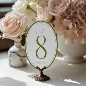 Puede incluir: Una tarjeta ovalada con el número de mesa 8 en verde, colocada sobre un soporte decorativo de bronce. La tarjeta está rodeada por un borde verde y presenta el texto "Grace & Oliver" y una fecha. El fondo incluye un arreglo floral con rosas.