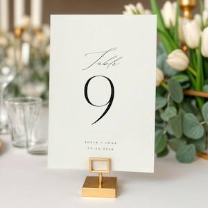 May include: A cream-colored table number card with the word "Table" and the number "9" in black script. The card is held in a gold-colored metal holder. The table is set with white linens, gold chargers, and glassware.
