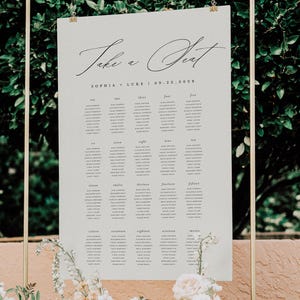 Könnte beinhalten: Ein Hochzeits-Sitzplan mit den Worten "Take a Seat" in eleganter Schrift. Die Namen der Gäste sind unten aufgeführt. Der Plan hängt an einem goldenen Rahmen und ist mit Blumen geschmückt.