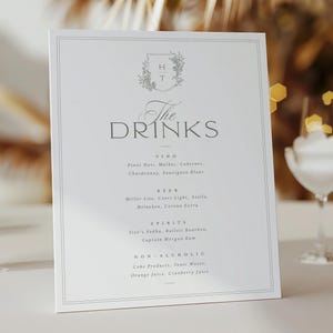 May include: A white rectangular menu card with the text "The DRINKS" in elegant script. The menu lists wine, beer, spirits, and non-alcoholic beverages. A lit candle in a glass cylinder and a wine glass are also visible.