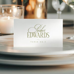 May include: A white place card with the text "Parker EDWARDS" in elegant gold script, and "TABLE NO. 1" in a smaller font. The card sits on a white plate, with candles and glassware in the background.