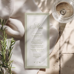 May include: A wedding menu card with a sage green border and elegant script. The menu lists starters, entrees, and dessert. A glass of water and sprigs of greenery are also in the image.