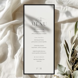May include: A formal menu card with a black frame, displaying a list of dishes in elegant script. The menu includes starters, main courses, and desserts, such as herb-roasted chicken and red velvet cake. The background is a white cloth.