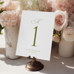 Puede incluir: Una tarjeta blanca con el número de mesa "1" en verde oliva, exhibida en un soporte de bronce. La tarjeta también incluye los nombres "Grace & Oliver" y la fecha "06.28.2028". La tarjeta se encuentra sobre un fondo de flores rosas y blancas.