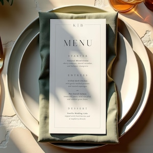 Op de afbeelding: Een formele tafelsetting met een wit bord, gouden vork en mes en een groene servet. Een menukaart met het woord "MENU" is gecentreerd op het bord. Twee glazen amberkleurige vloeistof staan in de rechterbovenhoek.