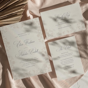 May include: A set of three cream-colored wedding stationery cards featuring elegant black script typography and a subtle, light-colored floral vine border. The cards include a main invitation, a details card, and an RSVP card with text.