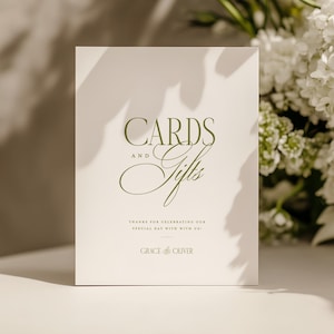 Könnte beinhalten: Eine weiße Karte mit den Worten "CARDS and Gifts" in eleganter grüner Schrift. Die Karte enthält auch den Text "THANKS FOR CELEBRATING OUR SPECIAL DAY WITH US!" und "GRACE & OLIVER". Weiße Blumen sind im Hintergrund.