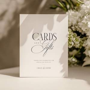 May include: A white card with the words "CARDS and Gifts" in elegant black script. The card also includes the text "THANKS FOR CELEBRATING OUR SPECIAL DAY WITH US!" and the names "GRACE & OLIVER". White flowers are in the background.