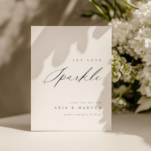 Puede incluir: Una tarjeta blanca con las palabras "Let Love Sparkle" en una elegante escritura. Los nombres "Aria & Marcus" están impresos debajo, junto con el texto "sparkler send off at 10:30 pm." Flores blancas en el fondo.