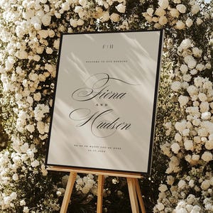 May include: A rectangular wedding welcome sign with a black frame on a wooden easel. The sign features the text F|H, Welcome to our wedding, Fiona and Hudson, We're so happy you're here, and the date 06.17.2026 in black serif and script fonts.