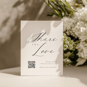 May include: A white card with the words "Share the Love" in elegant script, accompanied by a QR code and instructions. The card is set against a backdrop of white flowers, creating a soft, romantic aesthetic.