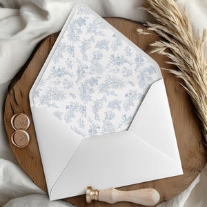 Pode incluir: Um envelope branco com forro estampado floral azul claro, exibido aberto sobre uma superfície de madeira. Também estão presentes dois selos de cera dourados com iniciais e um carimbo de cera com cabo de madeira. Um raminho de capim seco.