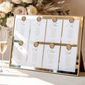 Könnte beinhalten: Ein goldgerahmter Spiegel-Sitzplan mit acht weißen Karten, die von One bis Eight in eleganter grüner Schrift beschriftet sind. Jede Karte ist mit einem goldenen Wachssiegel befestigt und listet Gästenamen auf. Links stehen zwei Sektgläser.