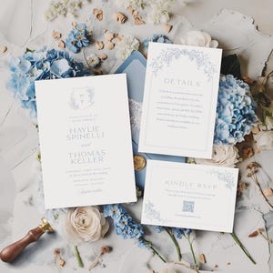 May include: Wedding stationery suite in pale blue and white, featuring an invitation, details card, and RSVP card, each with floral designs. The arrangement is set amongst blue hydrangeas and other flowers.