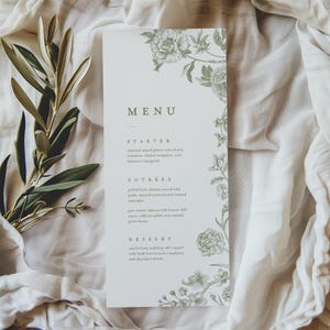 May include: A rectangular menu card with a floral design in green. The menu includes sections for starter, entrees, and dessert. The text is in a serif font. The card is on a white cloth.