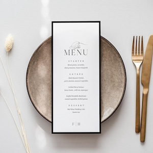 May include: A tall, rectangular white menu card with a thin black border, featuring the text THE MENU, STARTER, ENTREE, and DESSERT. The menu is centered on a round, speckled brown ceramic plate, accompanied by a gold-toned metal fork and knife.