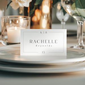 Puede incluir: Una tarjeta de sitio blanca con el nombre "Rachelle Reynolds" y el número "13" impresos en negro. La tarjeta está sobre un plato blanco, con velas y cristalería en el fondo. La tarjeta también tiene las iniciales "KIB" en la parte superior.