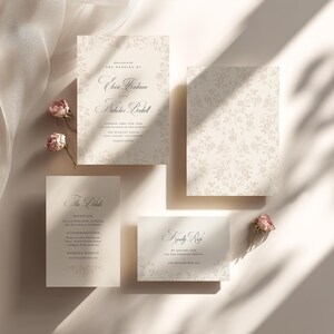 May include: A wedding stationery set featuring a main invitation, a reception card, and an RSVP card in cream with dark gray serif typography and floral borders. The set includes small dried pink rosebuds and a sheer white fabric accent.
