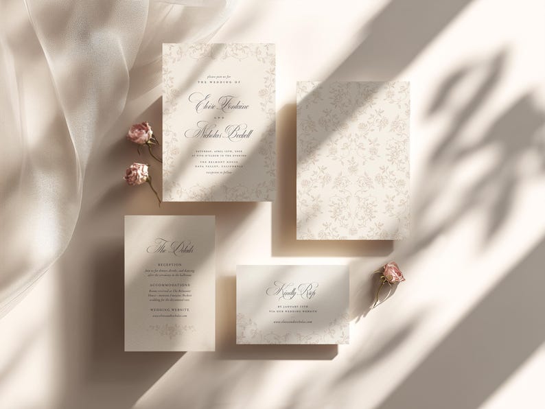 May include: A wedding stationery set featuring a main invitation, a reception card, and an RSVP card in cream with dark gray serif typography and floral borders. The set includes small dried pink rosebuds and a sheer white fabric accent.