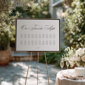 May include: A rectangular wedding seating chart sign with a black frame, featuring the text "Our Favorite People" in a black script font at the top, followed by a grid of twenty numbered tables with guest names and the initials "F|H" at the bottom.