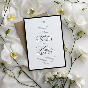 May include: Wedding invitation with a black border, featuring the names Fiona Bennett and Hudson Prescott. The invitation is surrounded by white orchids and a tan envelope, set against a white, silky background.