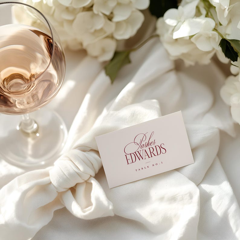 Puede incluir: Una copa de vino rosado junto a una tarjeta de mesa con el nombre "Parker EDWARDS" en escritura burdeos. La tarjeta est&aacute; sobre una tela blanca, con flores blancas al fondo. Las palabras "TABLE NO. 1" est&aacute;n impresas debajo.