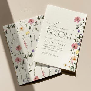 May include: Two white floral-themed cards. One card features the text "Love in Bloom" and event details. The other card has a floral pattern. Pink, yellow, and purple flowers are visible. The cards are on a light beige surface.