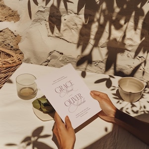 Puede incluir: Una invitaci&oacute;n de boda con los nombres "Grace Atwood" y "Oliver Sembriche" en escritura burdeos. La invitaci&oacute;n es sostenida por manos, con un vaso, un plato peque&ntilde;o con hojas y una taza visibles. El fondo incluye una pared texturizada y sombras.
