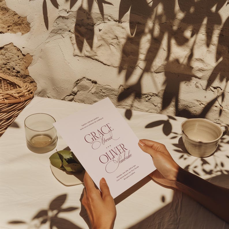 Puede incluir: Una invitaci&oacute;n de boda con los nombres "Grace Atwood" y "Oliver Sembriche" en escritura burdeos. La invitaci&oacute;n es sostenida por manos, con un vaso, un plato peque&ntilde;o con hojas y una taza visibles. El fondo incluye una pared texturizada y sombras.