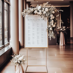 May include: A wedding seating chart with the names "Kayla and Blake" displayed on a white background, framed in gold. The chart is adorned with white roses and greenery. The stand is gold and rectangular. A bouquet of white roses sits at the base.