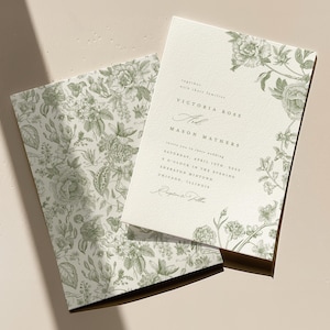 May include: A cream-coloured wedding invitation and matching envelope liner featuring a sage green botanical floral print. The invitation displays the text Victoria Rose and Mason Mathers, including the date Saturday, April 15th, 2023.