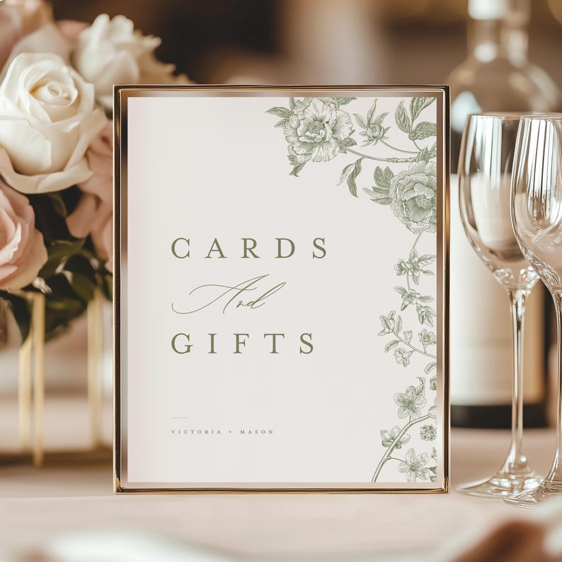 May include: A rectangular sign in a metallic gold frame featuring the text CARDS And GIFTS and the names VICTORIA + MASON in dark green font. The sign has a white background with a detailed botanical illustration of green flowers and leaves.